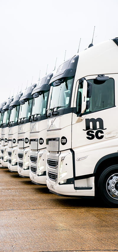 Fleet of MSC trucks parked in a row. The trucks are branded with the MSC logo and are set against a backdrop of a depot. The scene conveys a sense of organised logistics and transportation efficiency.